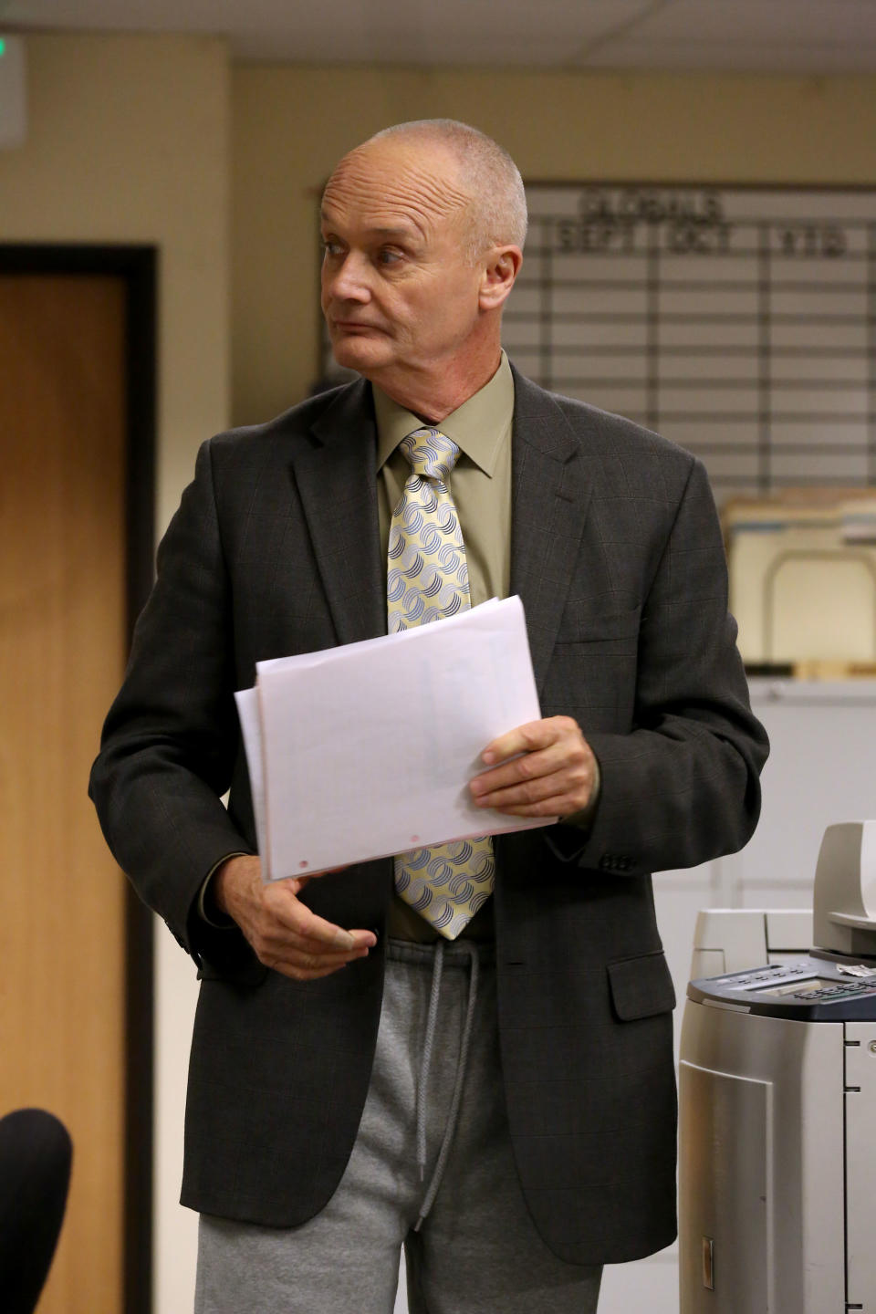 Creed Bratton wearing a suit jacket, tie, and sweatpants, holding papers, looking sideways in an office setting