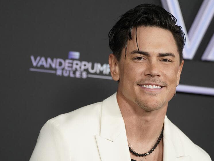 A man with dark hair and a dark beaded necklace wearing a cream colored suit jacket