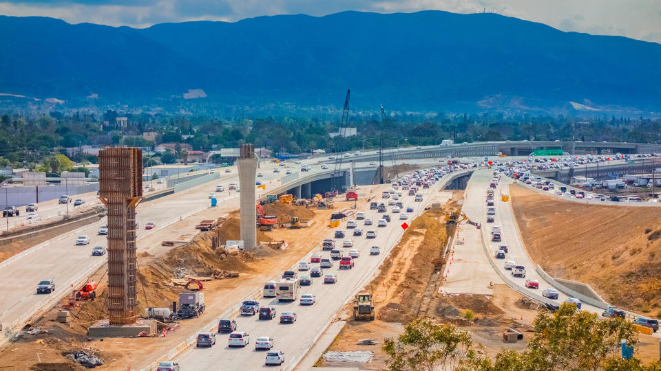 Overpass construction and equipment with traffic on the 91 Freeway fills the foreground leading back to hills in background with clouds above, Riverside California.