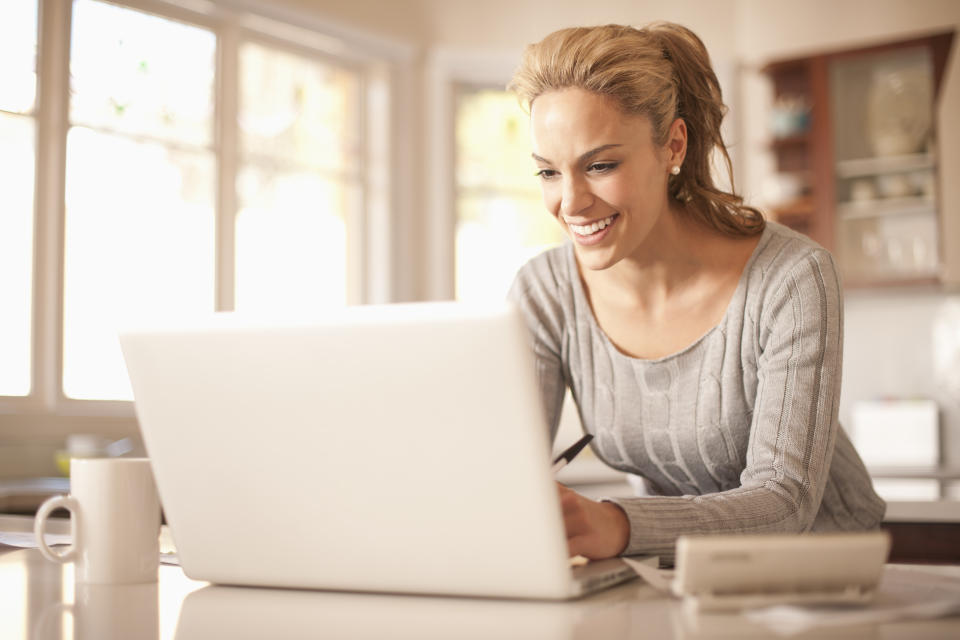 A woman smiles while organising her money on a laptop at her kitchen counter.