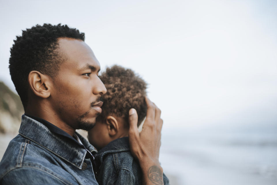 Man holds a child close to him as they both look away from the camera, standing near a body of water