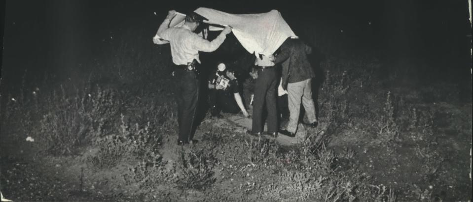 In this 1971 archival photo, police and detectives — protected from the rain — take footprint impressions on a path in the field where the body of Terri Lee Erdmann, 15, was found. She had been stabbed to death.