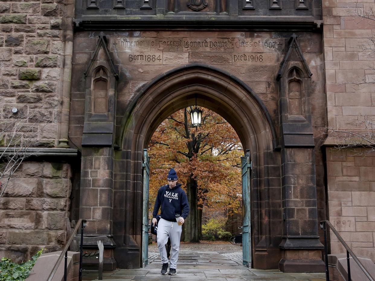 A student wearing a sweatshirt and sweatpants walks below a brick archway, with orange and yellow tree leaves in the background.