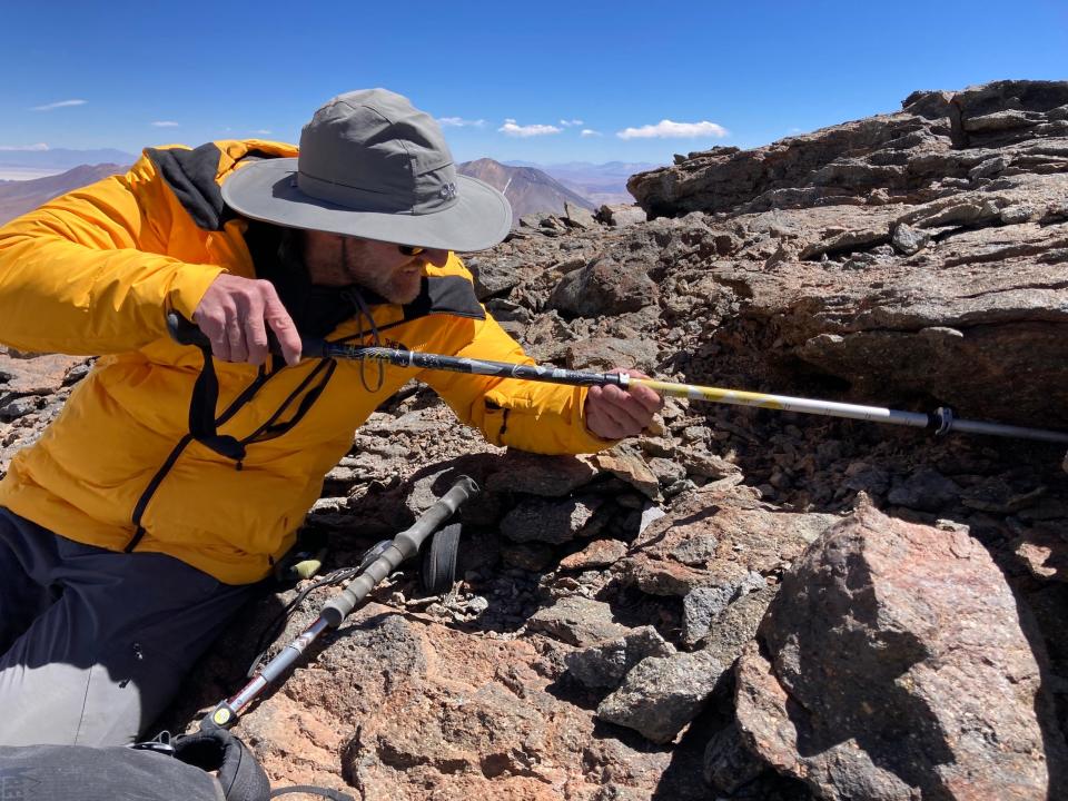 man in yellow windbreaker jacket and grey wide brimmed hat reaches a trekking pole into a crevice under a rock outcropping atop a barren mountain landscape