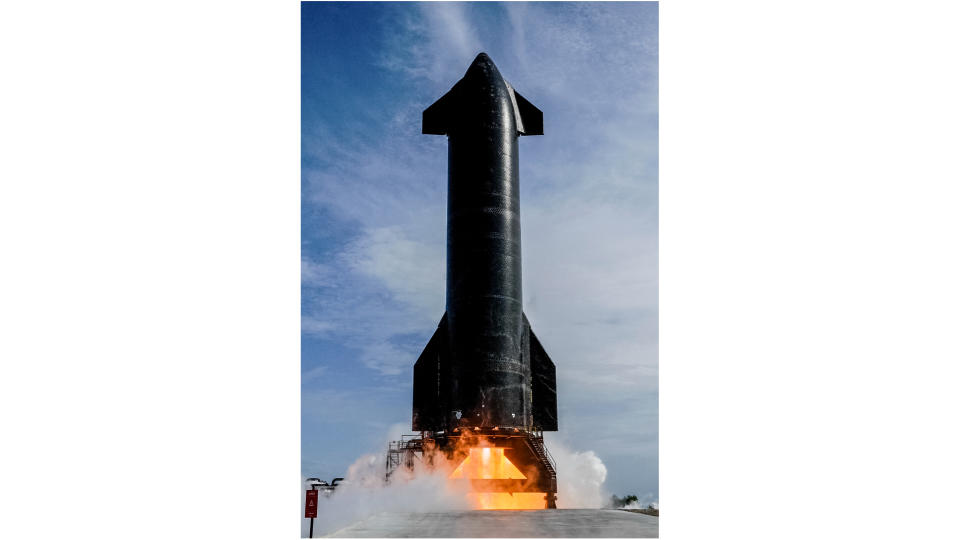 Eine große schwarze Rakete zündet ihre Triebwerke auf einem Außenprüfstand, im Hintergrund blauer Himmel