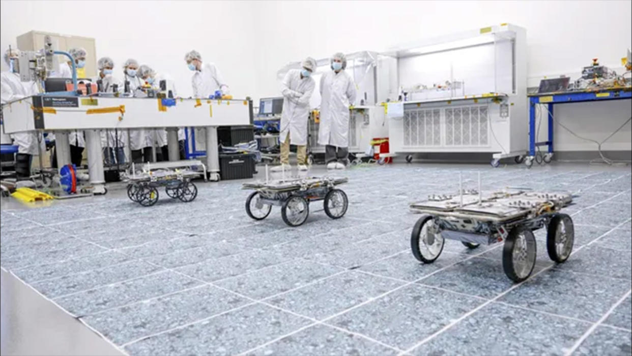  Three small rovers with four wheels each pictured in a lab with scientists standing in the background to observe. 