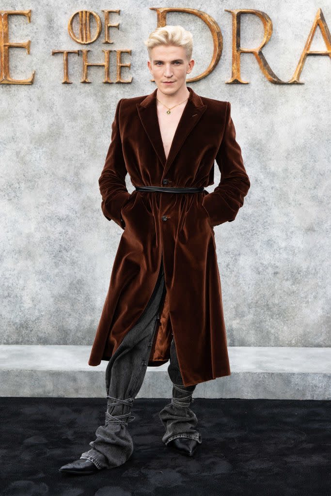 Tom Glynn-Carney on a grey carpet, wearing a deep brown velvet trench coat, grey trousers and boots, poses in front of "House of the Dragon" backdrop