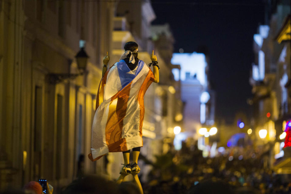 In this Wednesday, July 17, 2019 photo, a demonstrator with a Puerto Rican flag draped over his shoulders balances on the hands of another during clashes in San Juan, Puerto Rico. Thousands of people marched to the governor's residence in San Juan on Wednesday chanting demands for Gov. Ricardo Rossello to resign after the leak of online chats that show him making misogynistic slurs and mocking his constituents. (AP Photo/Dennis M. Rivera Pichardo)