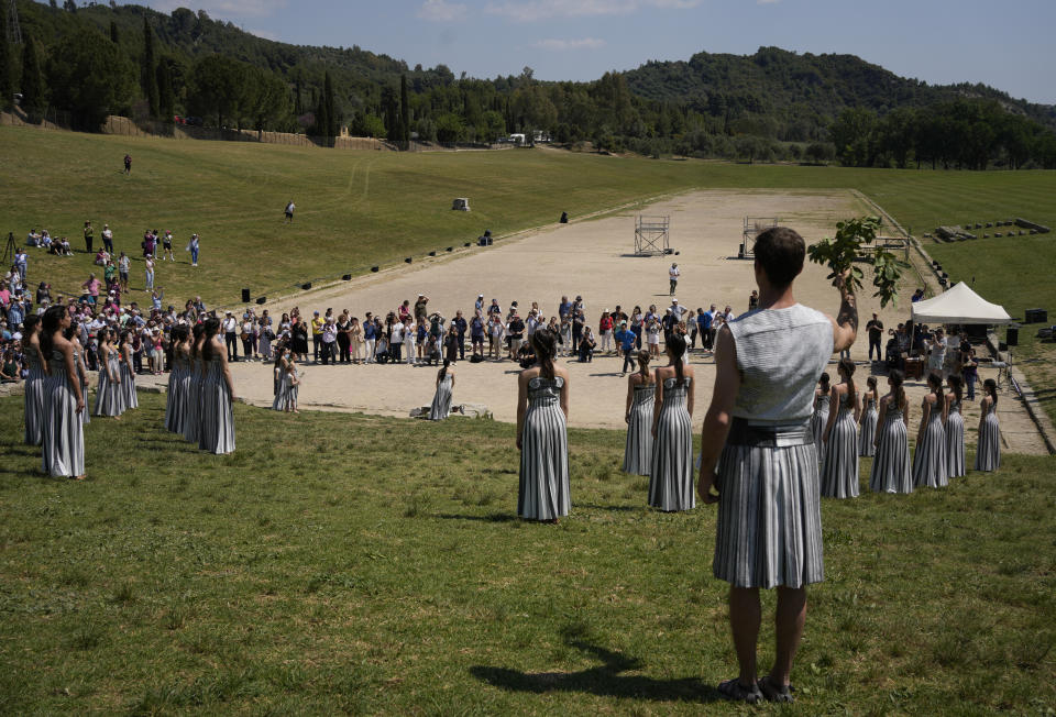 Visitors watch the performers, who will take part in the flame lighting ceremony for the Paris Olympics, during a rehearsal at Ancient Olympia site, Greece, Sunday, April 14, 2024. Every two years, a countdown to the Olympic games is launched from its ancient birthplace with a flame lighting ceremony in southern Greece at Ancient Olympia. The event is marked with a performance by dancers who assume the role of priestesses and male companions, their movement inspired by scenes on millennia-old artwork. (AP Photo/Thanassis Stavrakis)