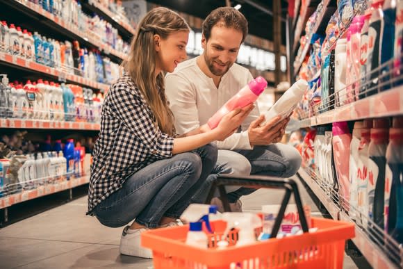 a young couple compares two household cleaners in a grocery store next to their basket full of items on the floor.