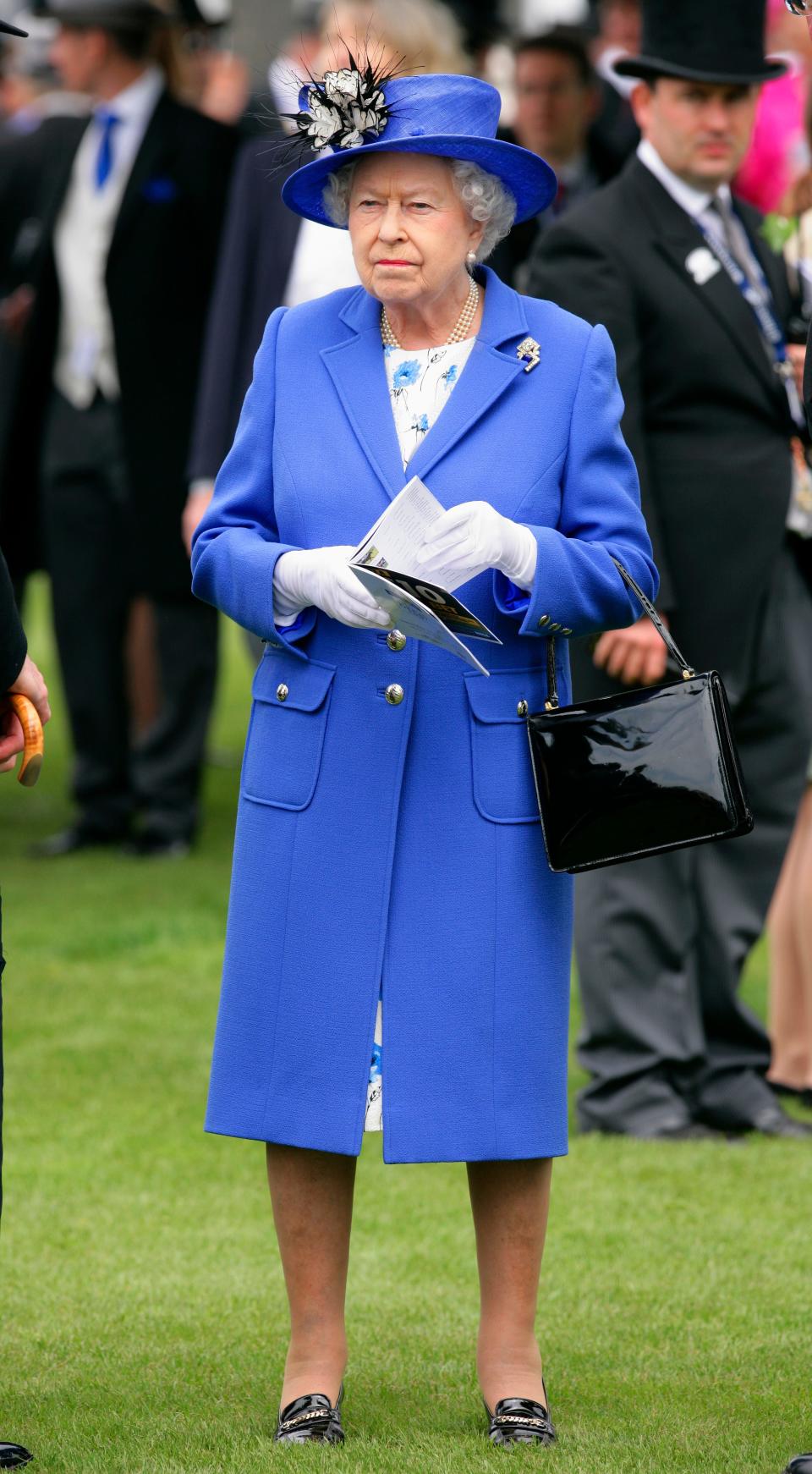 The Queen stands in a bright bluish-periwinkle overcoat with a v-shape neckline and collars revealing a white dress with a blue floral print on it. She's wearing a matching blue hat with a black and white flower on it. She has white gloves, a black shiny purse, and black flat shoes on.