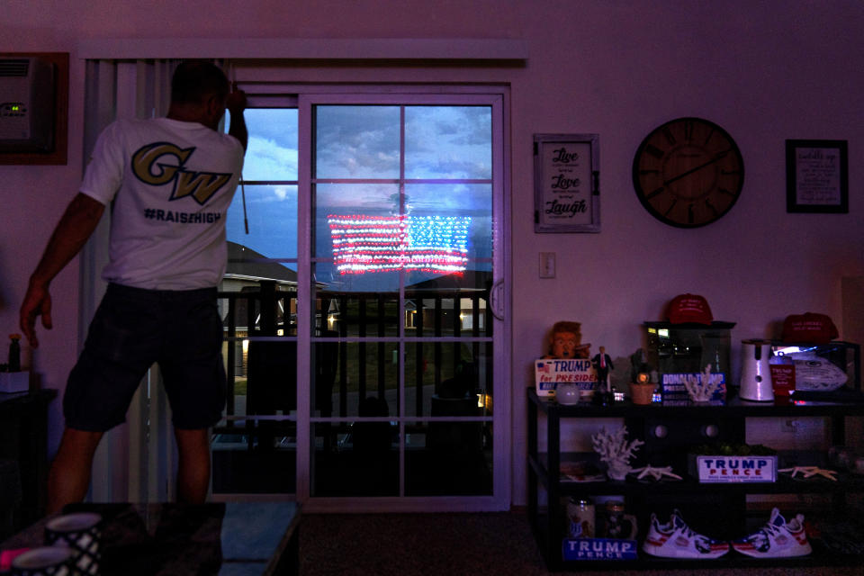 An illuminated American flag is reflected in the balcony door of Scott Rice's apartment in Appleton, Wis., Aug. 20, 2020. Rice reveres the president the way Wisconsin loves the Green Bay Packers. He has painted "T-R-U-M-P" on his lawn, spelled it out with Christmas lights on his roof and painted it on his steel-toed shoes. (AP Photo/David Goldman)