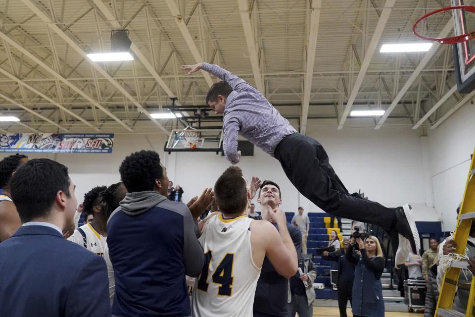 In this Thursday, Feb. 27, 2020 photo, Merrimack College head coach Joe Gallo leaps from a ladder into his players' arms after cutting down the net following a win over Central Connecticut in an NCAA college basketball game to claim a share of the Northeast Conference regular season championship in North Andover, Mass. The Warriors have been one of the biggest surprises in college basketball, winning more games than any other first-year Division I program in history. (AP Photo/Mary Schwalm)