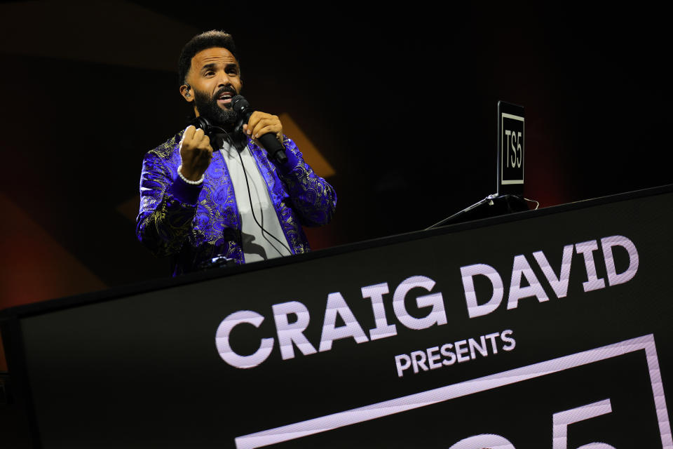 Craig David performing on stage, wearing a patterned blazer, holding a microphone, with a sign that reads "CRAIG DAVID PRESENTS" in front of him