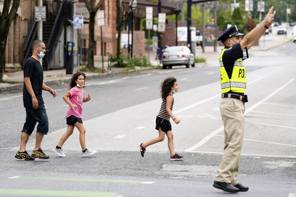 In this Sept. 9, 2020, photo Pedestrians cross an intersection as a cadet in the Baltimore Police Academy directs traffic during an on the field class session in Baltimore. (AP Photo/Julio Cortez)