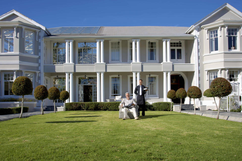 A man in a light suit sits on a garden chair in front of a large mansion, with another man standing beside him in a uniform