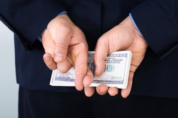 A man in a suit holding a stack of hundred dollar bills behind his back while also crossing his fingers.