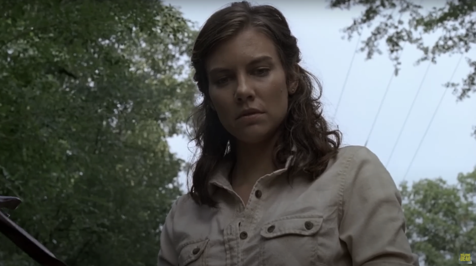 Lauren Cohan in a forest looking down with a serious expression. She wears a buttoned-up shirt