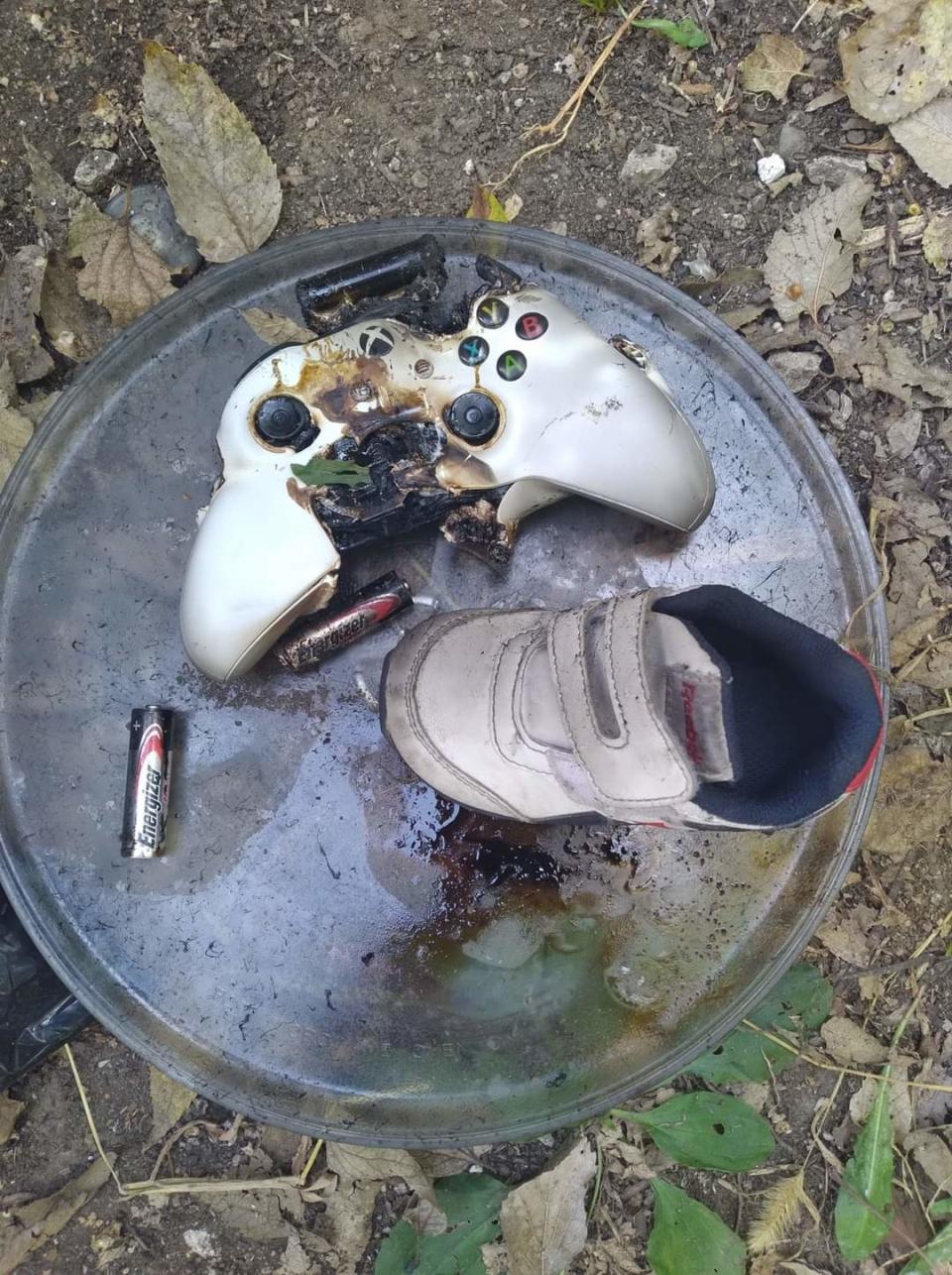 Damaged video game controller with melted parts and a small shoe on a metal surface, surrounded by batteries and dry leaves