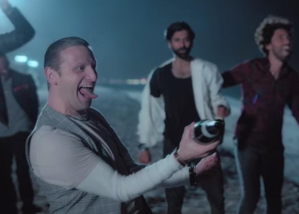 man wears a vest over a henley and pops a champagne bottle on the beach.