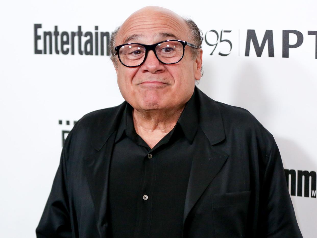 danny devito at a red carpet event, wearing a black shirt, black jacket, and black frame glasses. he's smiling slightly and has his eyebrows raised