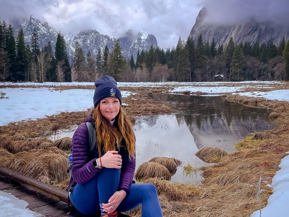 Emily sits on a wooden path covered in ice in Yosemite National Park. Behind her are mountains, trees, and a swampy area covered in snow.