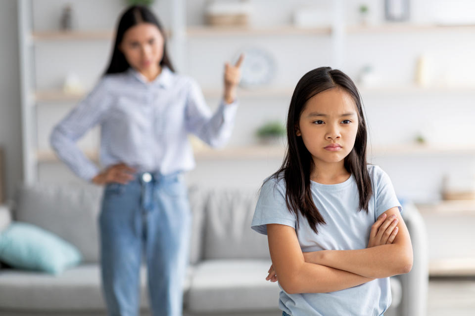 A girl with crossed arms looking upset with a woman gesturing in the background inside a home