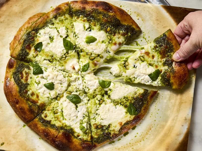 overhead shot of a full pesto pizza, on parchment paper, topped with basil leaves, with a hand reaching in and grabbing a slice