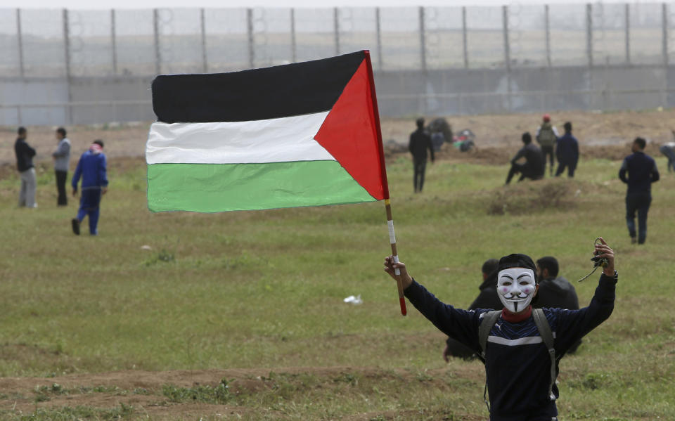 A masked protester waves his national flag near the fence of the Gaza Strip border with Israel, marking first anniversary of Gaza border protests east of Gaza City, Saturday, March 30, 2019. Tens of thousands of Palestinians gathered Saturday at rallying points near the Israeli border to mark the first anniversary of weekly protests in the Gaza Strip, as Israeli troops fired tear gas and opened fire at small crowds of activists who approached the border fence. (AP Photo/Adel Hana)