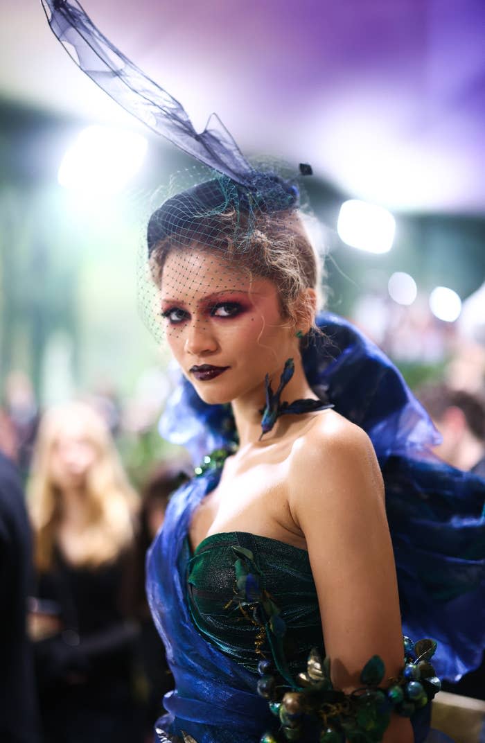 Zendaya in a blue-themed elaborate costume with a hat and veil; makeup includes red eyeshadow