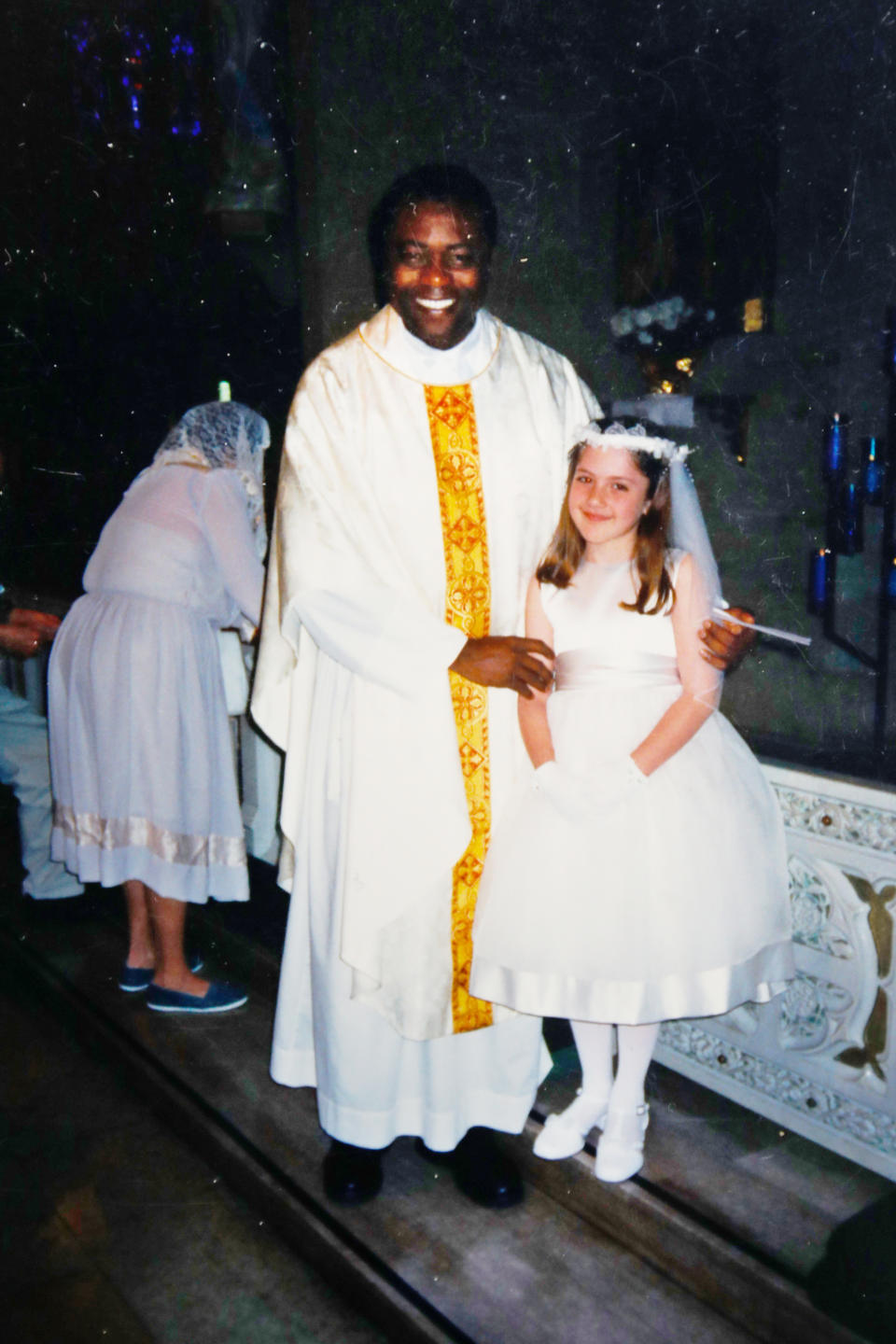 This undated photo provided by Mary Rose Maher in 2019 shows her as a child standing with Rev. Komlan Dem Houndjame at The Assumption of the Blessed Virgin Mary Church in Detroit. Two years after arriving at The Assumption of the Blessed Virgin Mary Church in 1999, Detroit Archdiocese officials said they asked Houndjame to return to his home country, Togo, after learning of accusations of sexual misconduct against him in Detroit and at an earlier posting in Florida. Instead he went to a treatment facility in St. Louis. In 2002, Detroit police arrested him and charged him with sexually assaulting a member of the church's choir. (Courtesy Mary Rose Maher via AP)