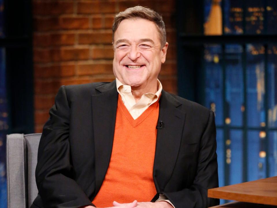 John Goodman waering an orange V-neck jumper and a black blazer while sitting on the set of "Late Night with Seth Meyers"