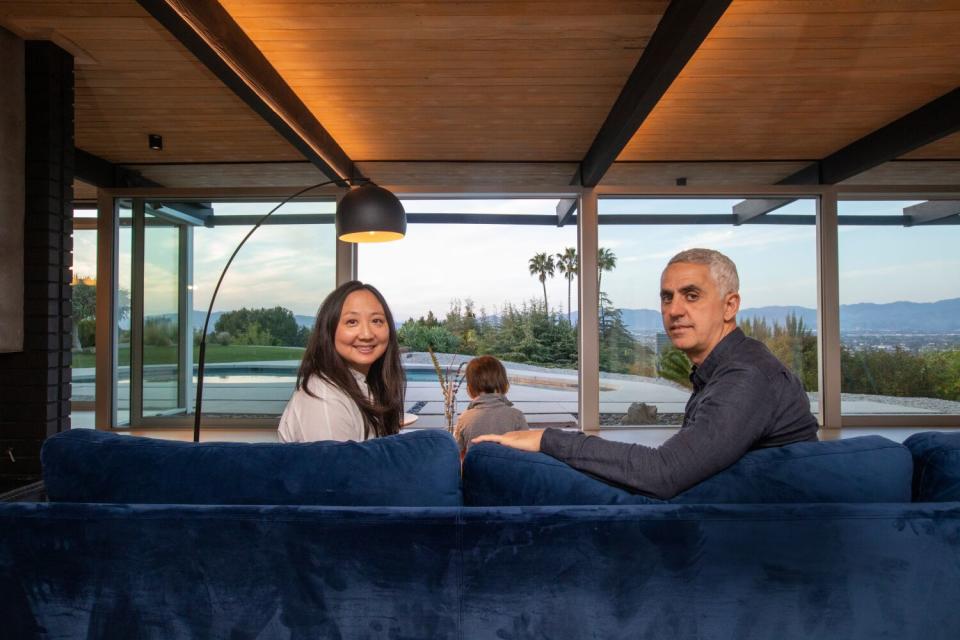 A couple seated on a sofa look over their shoulders at the camera, a boy and glass windows with a great view before them