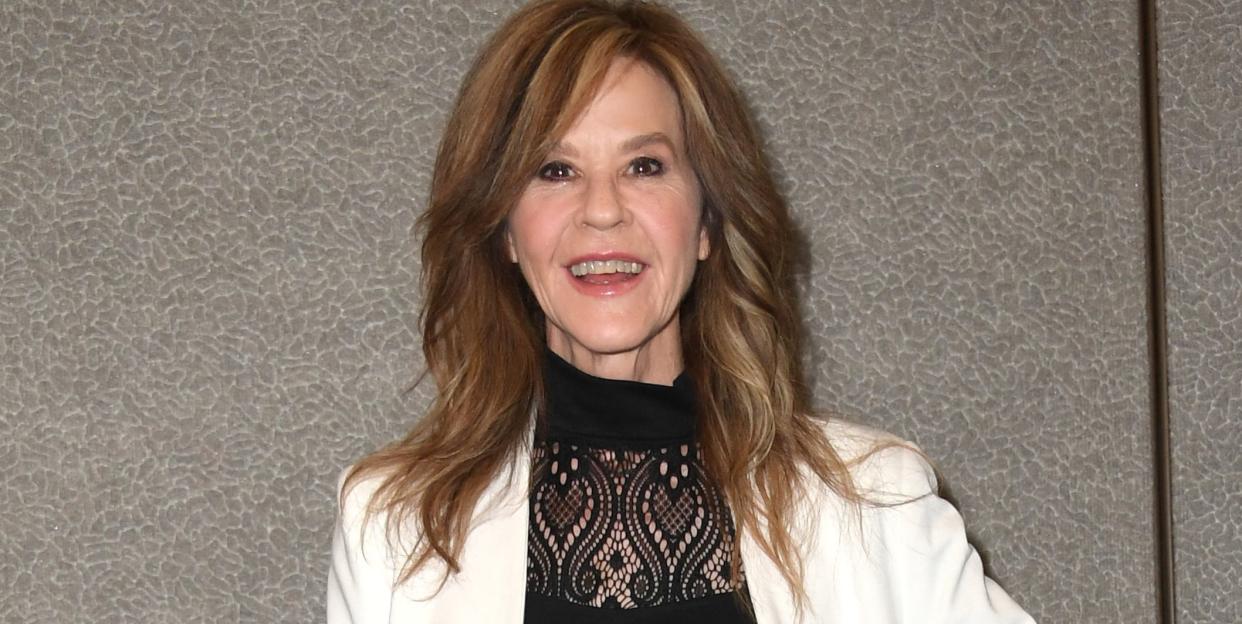 linda blair, a mature woman stands smiling and looking at the camera, she has shoulder length brown hair worn in loose waves, she wears a black top with black trousers and a white jacket