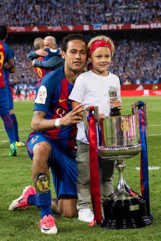 neymar jr and his son davi pose for a photo with a large trophy on a soccer field, davi stands next to the trophy and neymar kneels on one knee behind him,