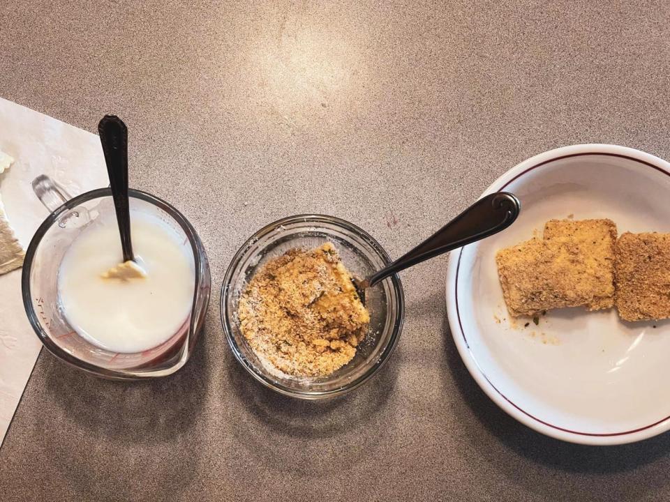 a cup of dredging liquid and a cup of breadcrumbs next to a plate of breaded ravioli