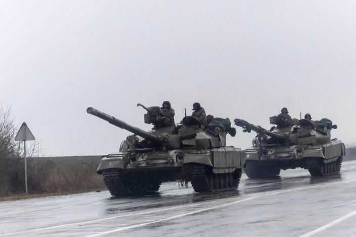 Two Ukrainian tanks, each with two servicemen visible on board, move down a rain-slicked road under gray skies. 