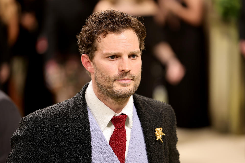 Jamie Dornan at a formal event, wearing a textured suit jacket, light-colored shirt, red tie, and gold pin