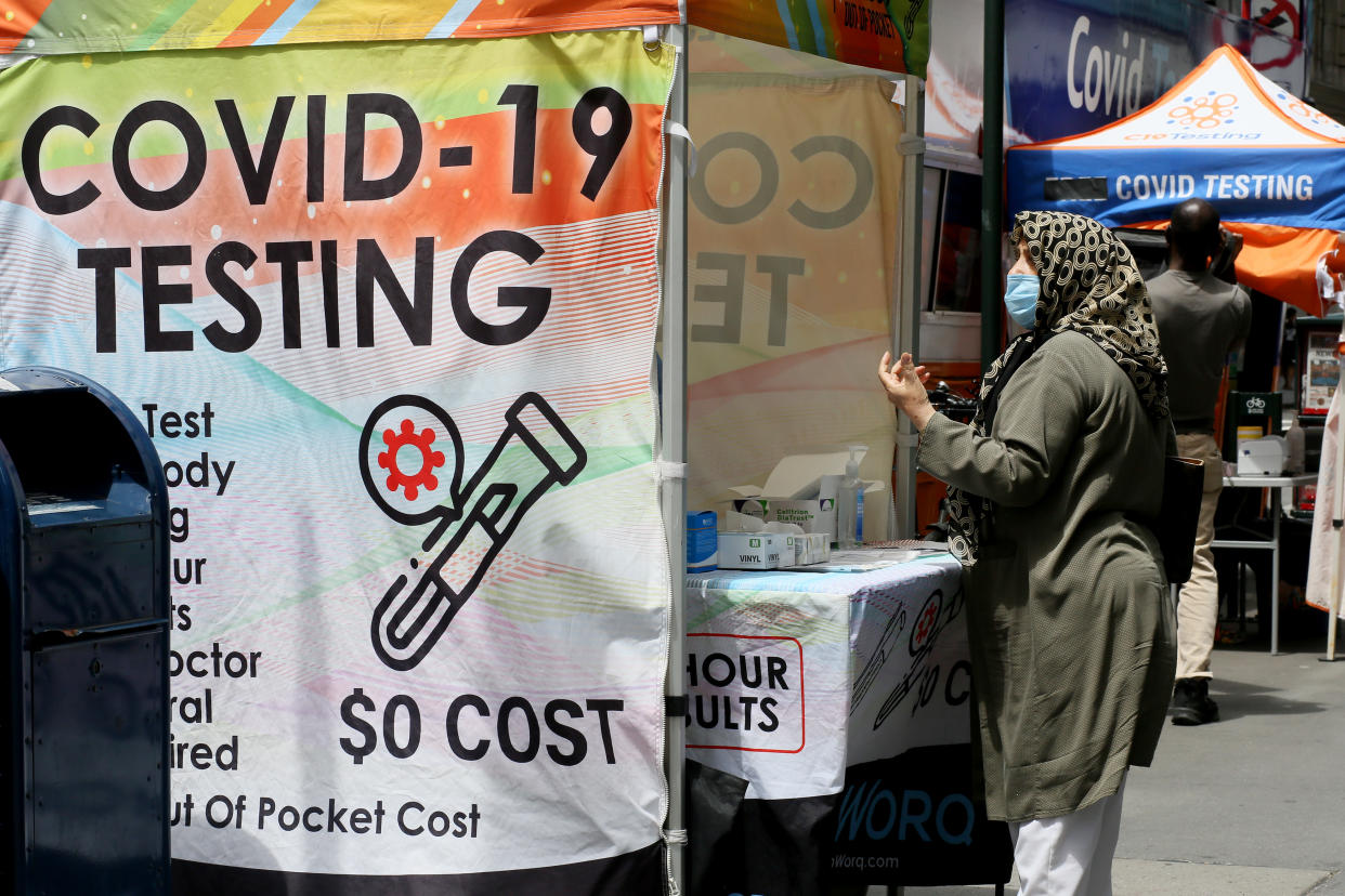 A woman in headscarf and mask waits at a booth saying: COVID-19 Testing, $0 Cost.