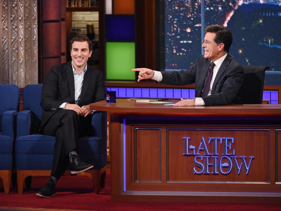 Chesky sit next to a wooden desk manned by Stephen Colbert