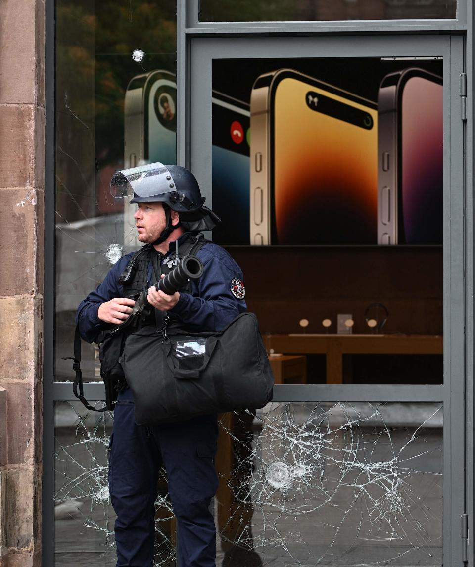 A French police officer in riot gear stands guard holding a tear gas canister launcher next to the facade of a damaged Apple Store at Place Kleber, in Strasbourg, eastern France, on 30 June 2023 (AFP via Getty Images)