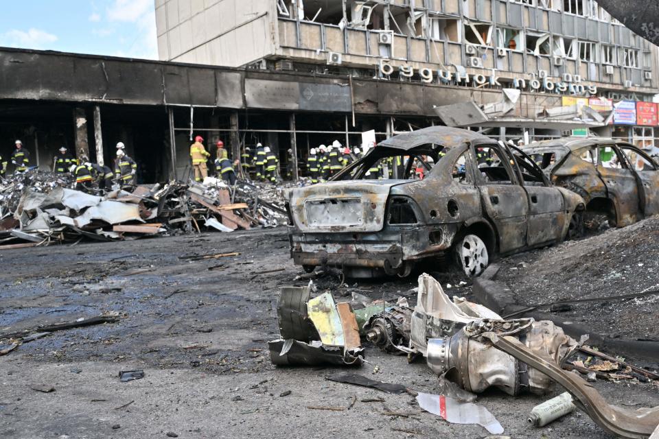 Rubble is seens along the sides of a street along with destroyed, burned cars and a damaged building.