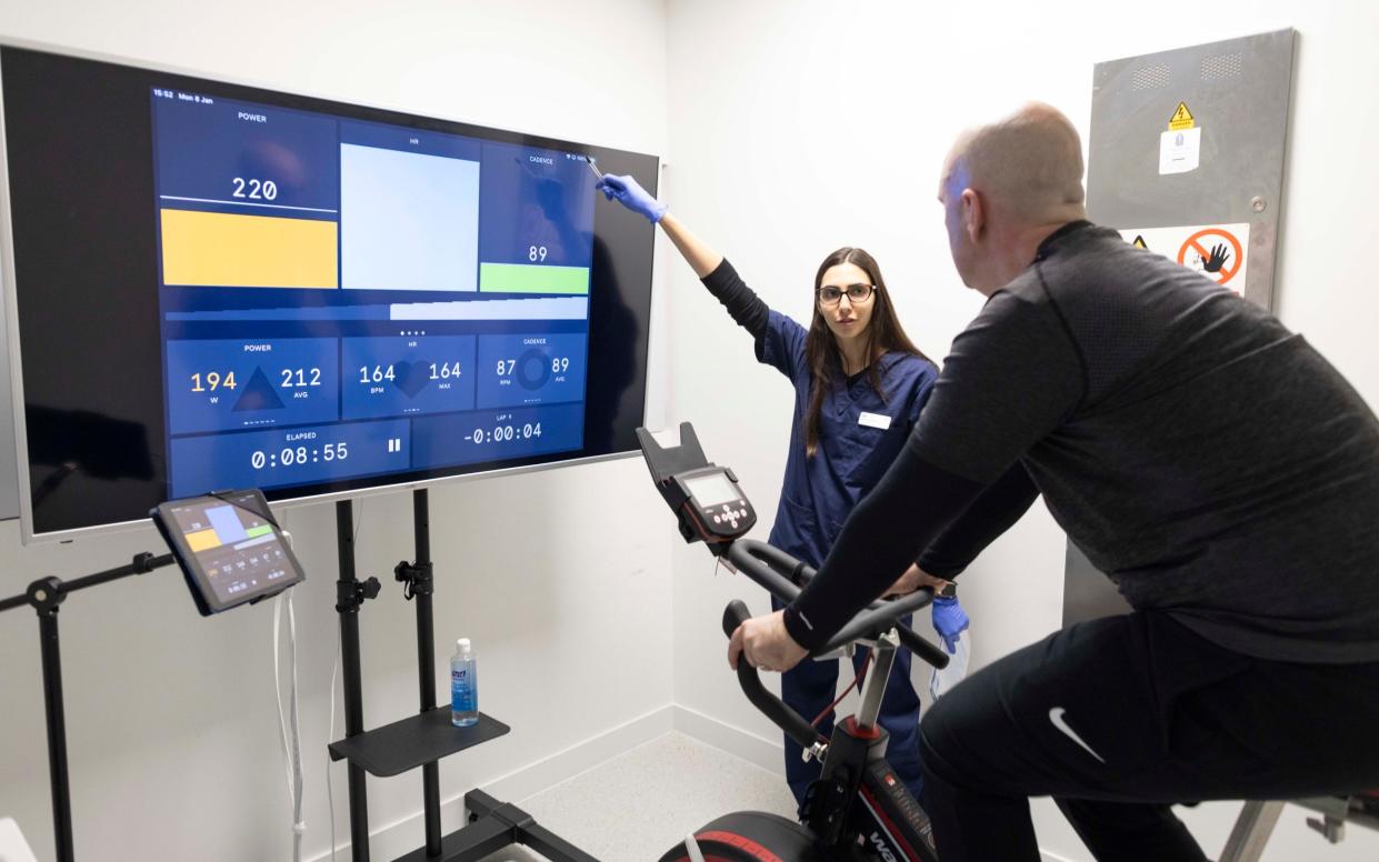 Sam's cardiovascular performance is displayed on a large screen in front of the watt bike