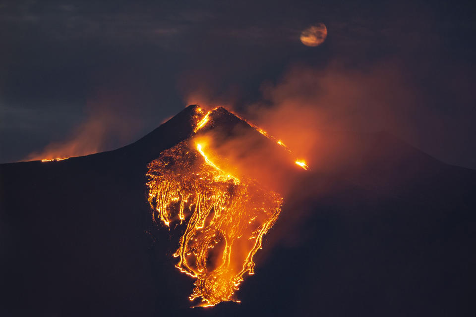 The moon is partially seen in the sky as lava flows from the Mt Etna volcano, near Catania in Sicily, southern Italy, early Tuesday, Feb. 23 , 2021. The explosion started before midnight on Monday night, provoking a huge eruption plume that rose for several kilometers from the top of Etna, as reported by The National Institute of Geophysics and Volcanology, Etneo Observatory. (AP Photo/Salvatore Allegra)