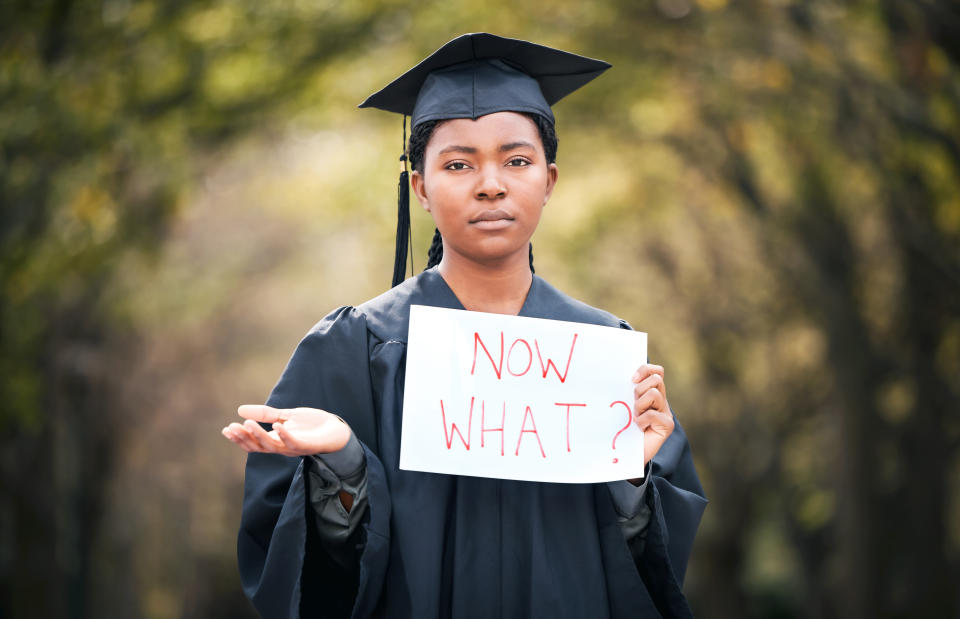 person in their graduation cap and gown with a sign that reads, now what?