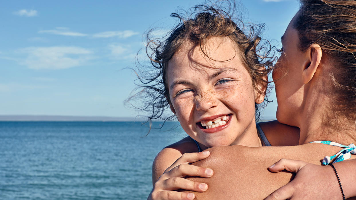  Portrait of girl with lots of freckles on her nose and cheeks hugging her mom on beach and smiling. 