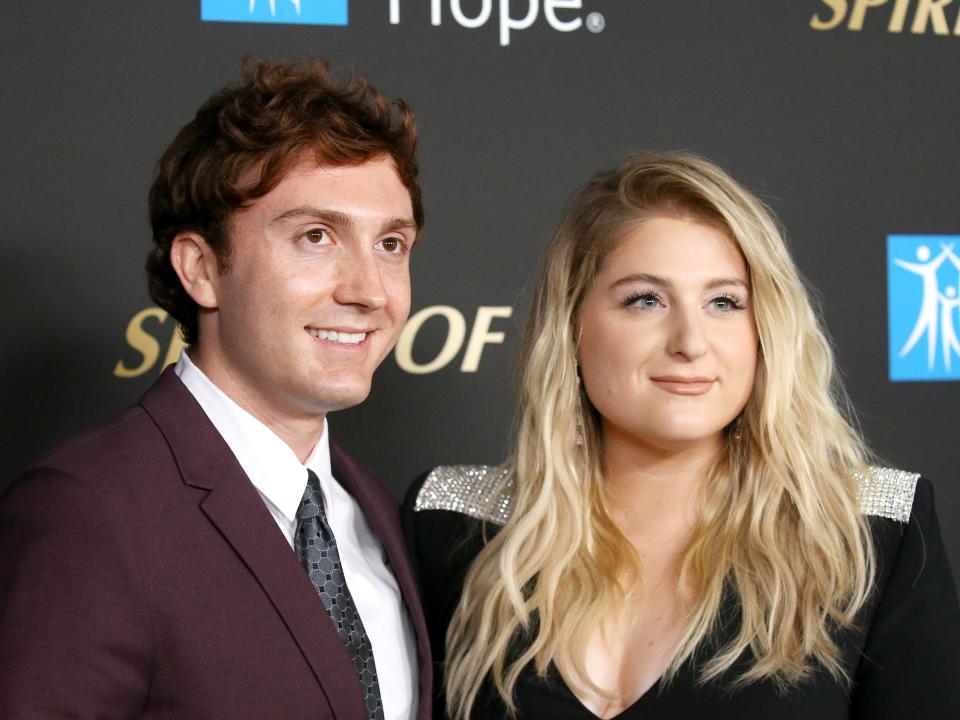 daryl sabara and meghan trainor on a red carpet, posing together and smiling. sabara is wearing a burgendy suit and grey tie, while trainor is in a black outfit with sparkling silver shoulders and her hair styled in waves