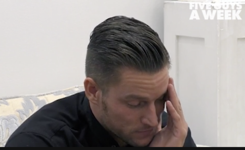 Man with slicked-back hair resting head in hand, looking distressed, in an indoor setting