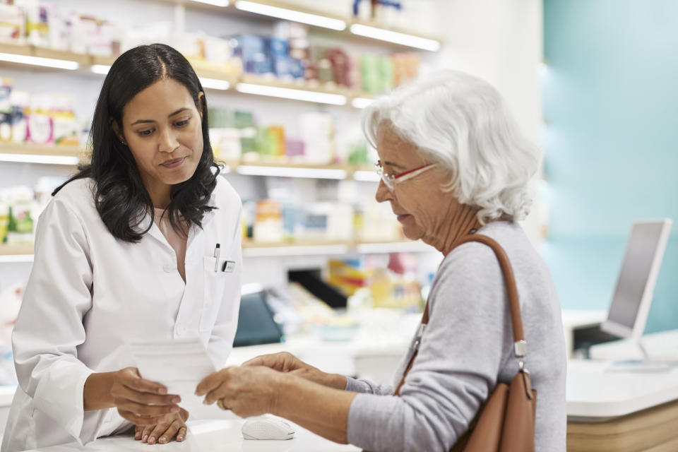 Senior customer showing prescription to female doctor. Cashier is assisting elderly woman at checkout counter. They are standing at pharmacy.