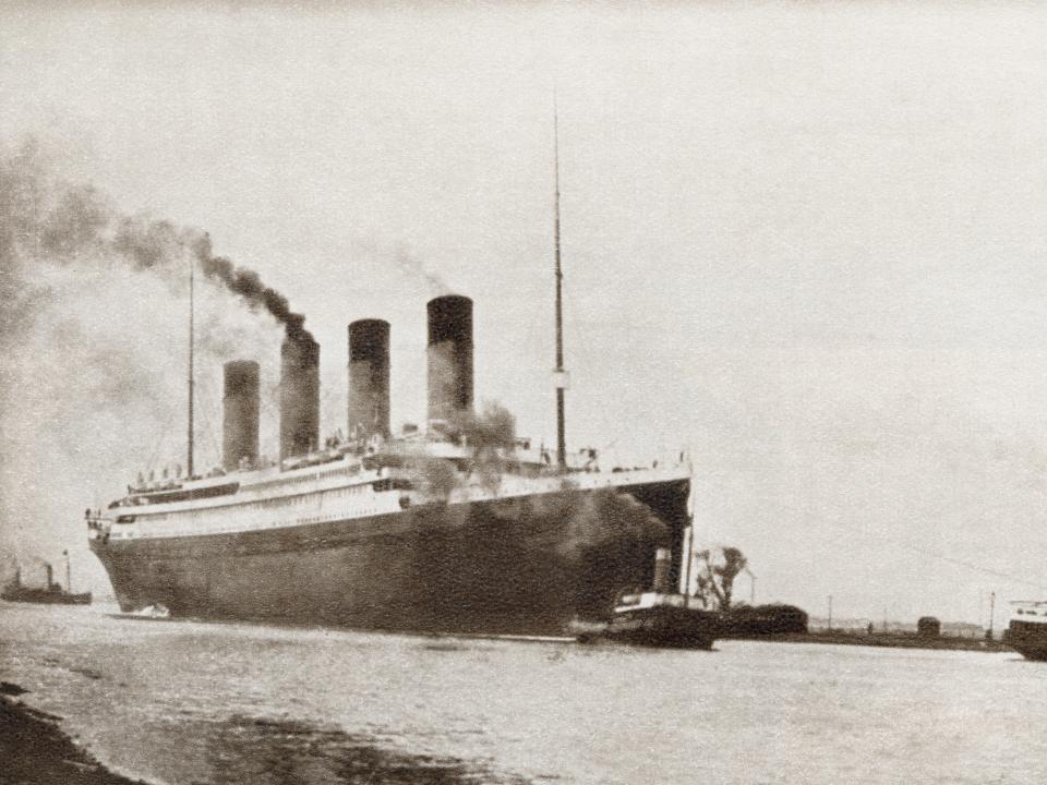 A black-and-white image of a smaller boat sailing in front of the Titanic, which has smoke stacks that are leaving a trail of soot in the air.